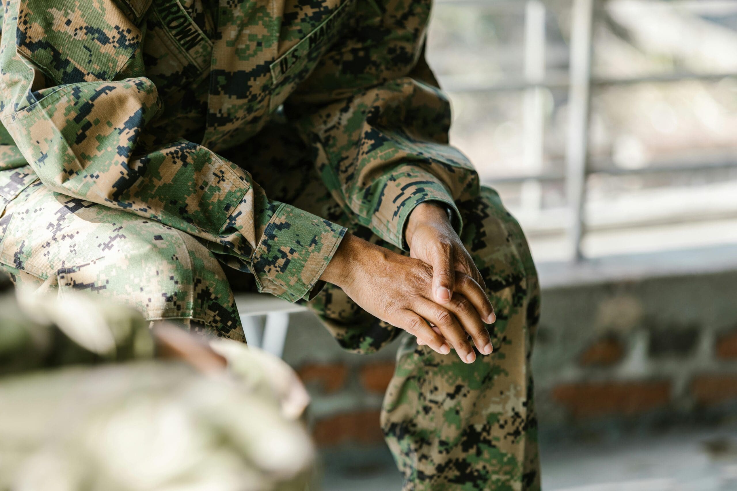A person wearing camouflage military uniform sitting with hands clasped, partially out of frame.